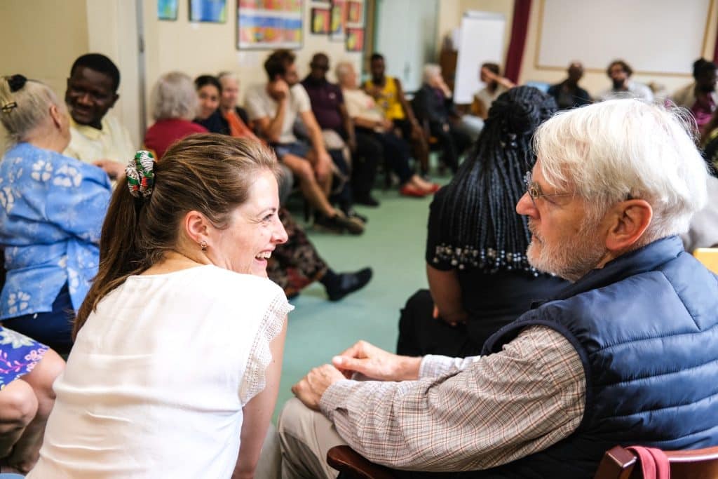 Une collaboratrice d’entreprise discute avec une personne âgée résidente du centre d’accueil Delta7 au premier plan, dans le cadre d’un exercice de théâtre d’improvisation pour un team building organisé par Lutila.